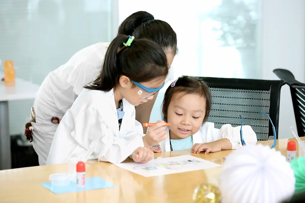 Two children and a woman in a research coat colour a picture at a table