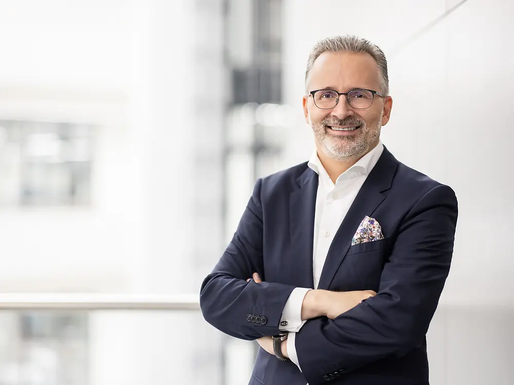 Carsten Knobel in a dark suit with a white shirt and pocket square, standing with arms crossed in a bright modern indoor setting.