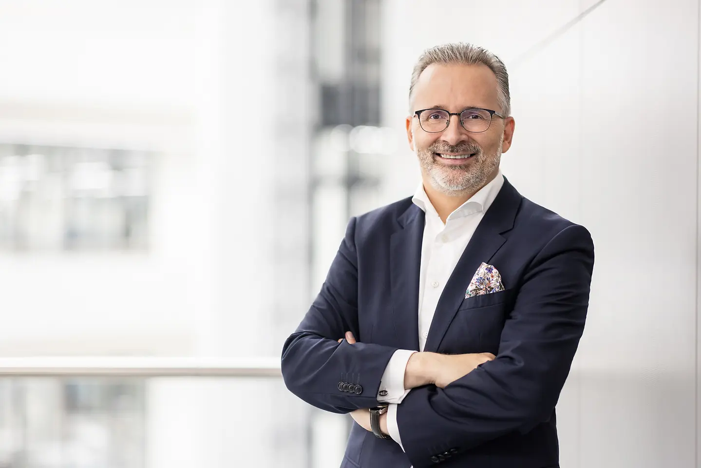 Carsten Knobel in a dark suit with a white shirt and pocket square, standing with arms crossed in a bright modern indoor setting.