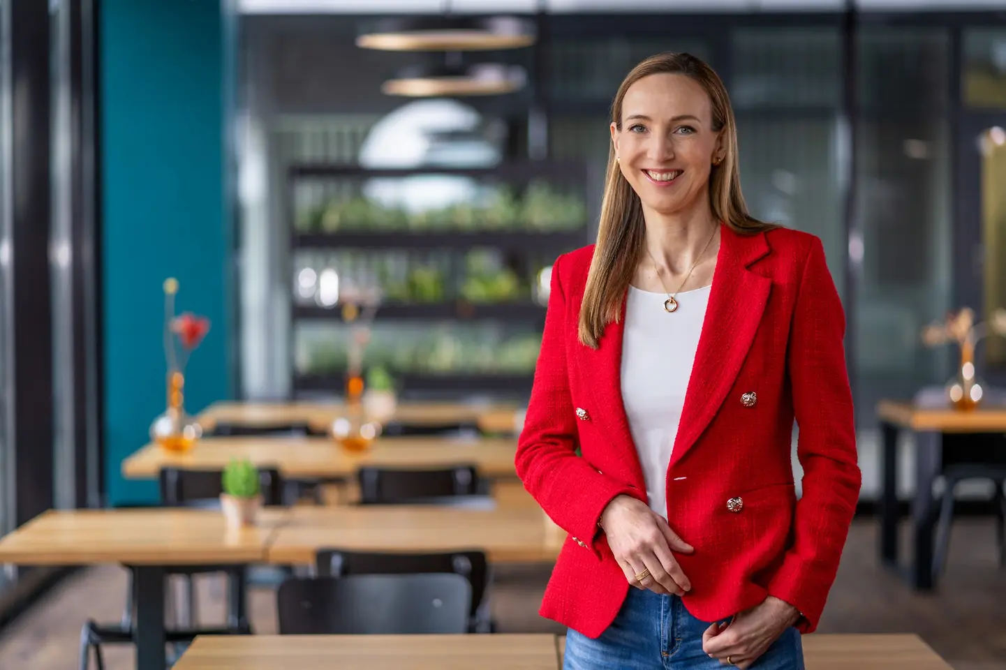 Dr. Simone Bagel-Trah wearing a bright red blazer and white top, standing in a modern café-style setting with wooden tables and decorative vases in the background.