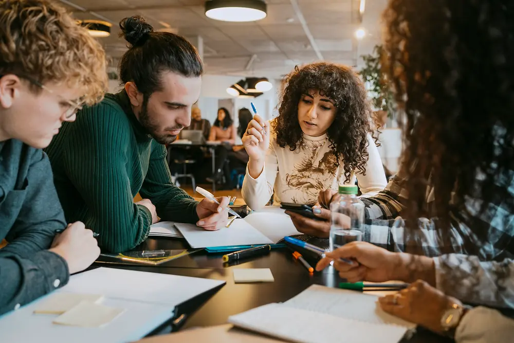 A group of people sitting together at a table in a modern indoor workspace, collaborating on documents, notebooks, and digital devices. Various pens, papers, and sticky notes are spread across the table while the group engages in discussion and note-taking.